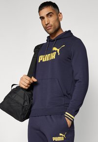 Felpa blu con logo giallo Puma, cordoncino e tasca frontale. Abbinata a pantaloni coordinati. Il soggetto tiene una borsa da viaggio nera.