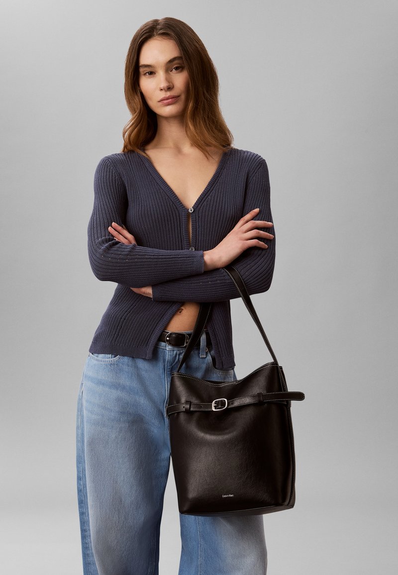 Femme aux longs cheveux bruns portant un cardigan côtelé foncé et un jean bleu clair, tenant un sac à main en cuir noir Calvin Klein.