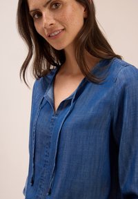 Une jeune femme avec des taches de rousseur légères et des cheveux bruns porte un blouse en denim bleu avec un décolleté en V et des liens à cordon.