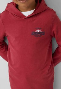 Kind in einem roten Kapuzenpullover mit weißem T-Shirt darunter, auf dem "Der Wilde Akrobatik-Zirkus"-Text und ein Zeltdesign auf der Brust zu sehen sind.