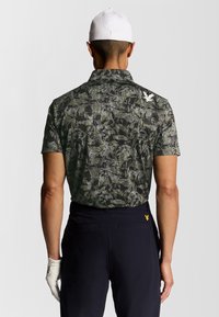 Manlig golfspelare som bär en vit keps, mönstrad mörkgrön pikétröja, svarta byxor med gult logo och en vit handske på vänster hand, visad bakifrån.