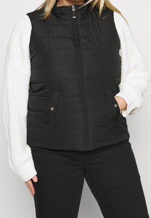 Kvinde iført en sort quiltet ærmeløs vest med lynlås og trykknaplommer over en hvid strikket sweater og sorte bukser.