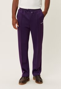 Pantalones deportivos morados ligeros con cintura ajustable con cordón, costuras laterales y un logo impreso en el muslo izquierdo. Textura suave.