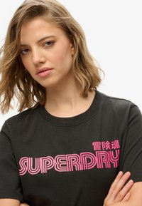 Czarna bawełniana koszulka z odważnym różowym napisem "SUPERDRY" z paskowymi akcentami. Klasyczny krój z okrągłym dekoltem.