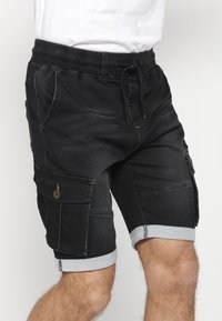 Pantalones cortos de cargo de denim negro con una cintura elástica, cordón, bolsillos laterales y puños doblados. Presenta costuras amarillas y detalles de botones.