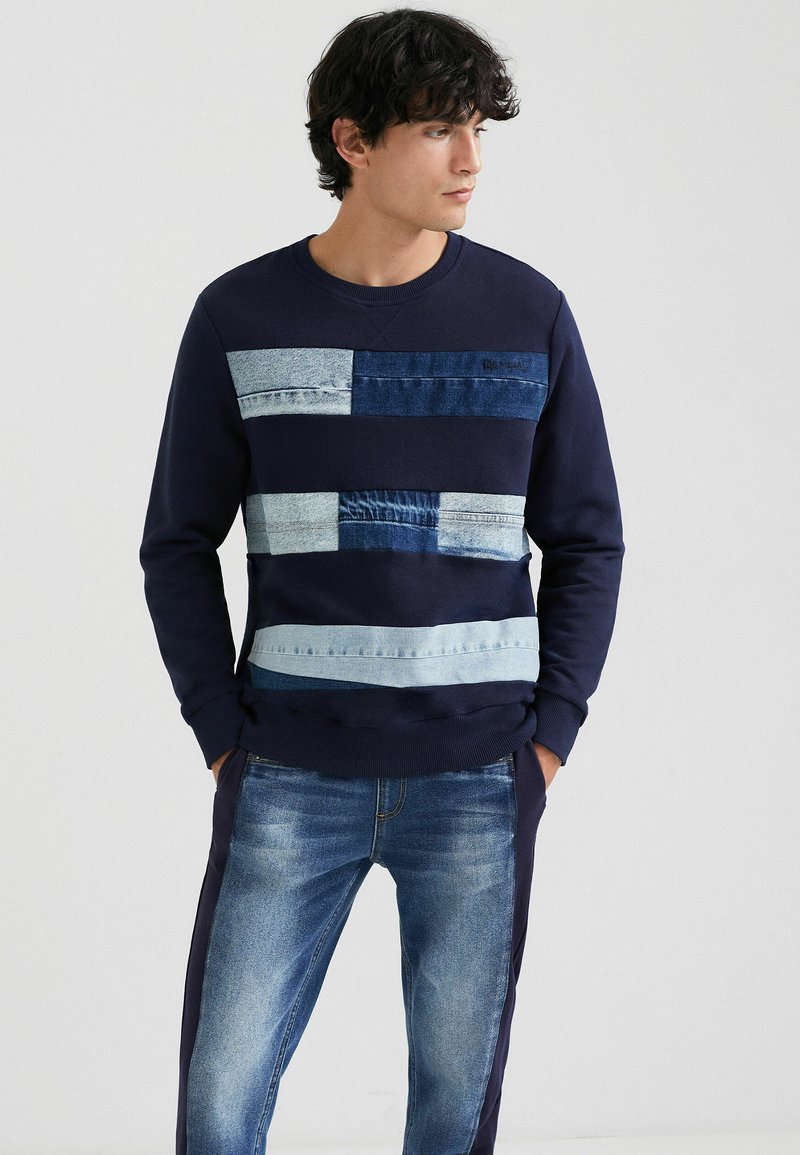 Sweatshirt navy avec un design en patchwork de différentes teintes de denim sur le devant. Le tissu est doux, avec des poignets et un col côtelés.