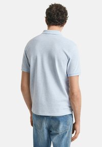 Man met krullend haar, gekleed in een lichtblauwe polo met korte mouwen en blauwe spijkerbroek, draaiend met de rug naar de camera, tegen een effen witte achtergrond.