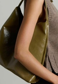 Olijfgroene leren tote bag met een zachte glans, grote vorm en enkele handvat, tegen een bruin gebreid top gehouden.