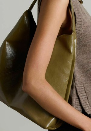 Olijfgroene leren tote bag met een zachte glans, grote vorm en enkele handvat, tegen een bruin gebreid top gehouden.