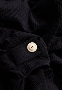 Tissu noir texturé avec un motif en tissage, présentant un bouton couleur crème et un boutonnière arrondie sur un détail de poignet.