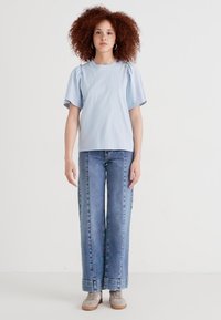 Haut bleu clair à manches courtes avec des épaules bouffantes, associé à un jean en denim clair à jambes larges. Des baskets grises avec une texture douce complètent la tenue.