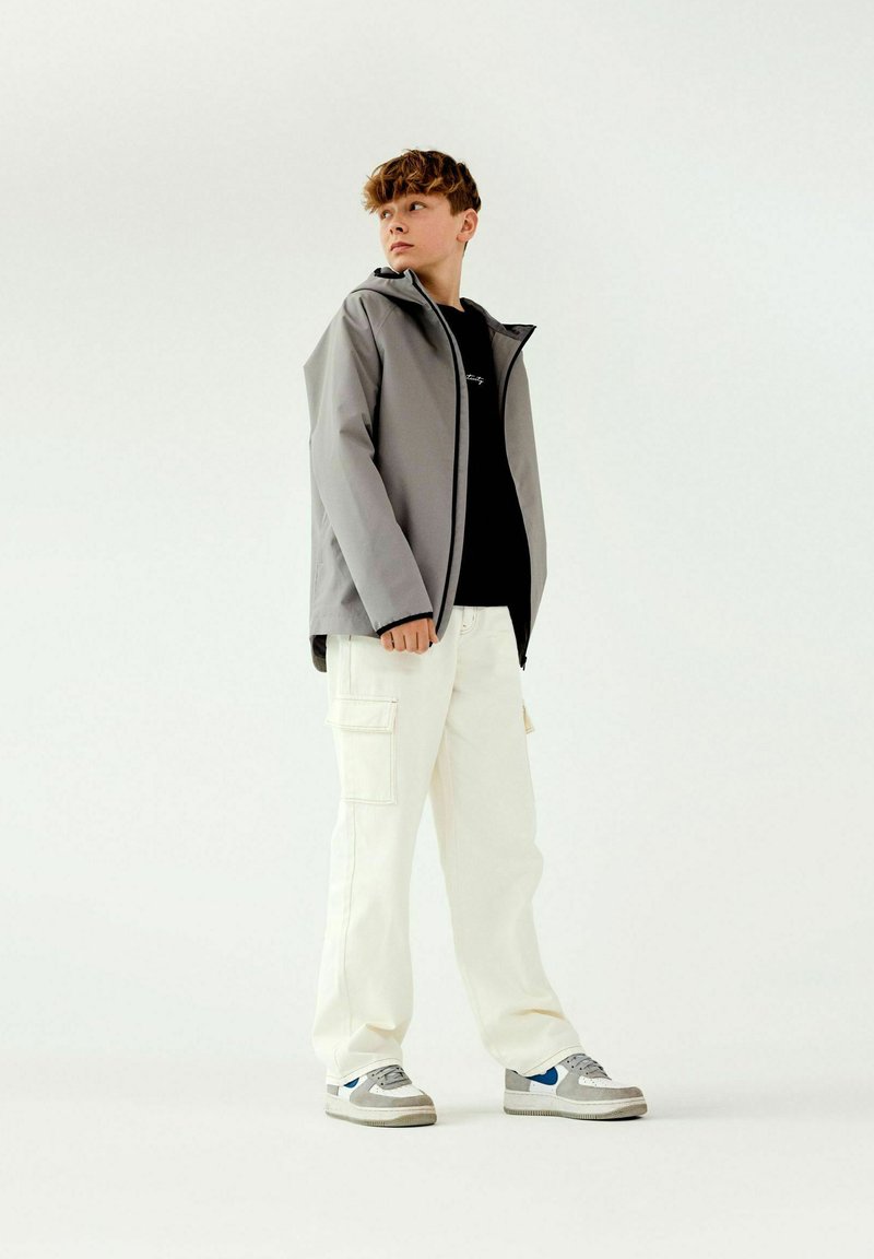 Veste imperméable grise avec capuche, portée par-dessus une chemise noire. Associée à un pantalon cargo blanc et des baskets grises avec des accents bleus.