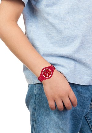 Montre-bracelet rouge avec un cadran rond, des chiffres blancs et un design ludique. Le bracelet présente un motif texturé, associé à une chemise bleu clair.