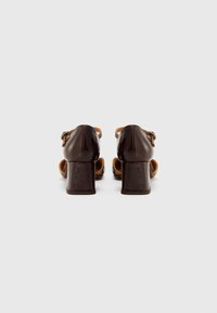 Chaussures à talons hauts en cuir verni marron avec bout rond, sangle cheville réglable et talon bloc texturé. La vue arrière montre des côtés ouverts.