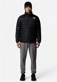 Čierna puffa bunda s horizontálnym prešívaním, oblečená cez červené tričko, doplnená sivými teplákmi a čiernymi topánkami. Model nosí čiernu čiapku.