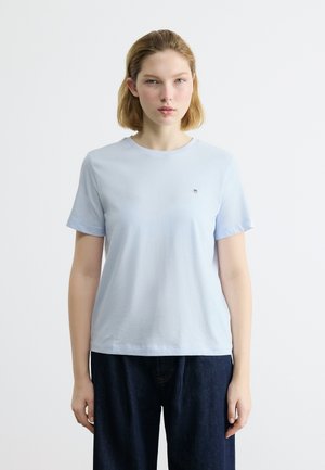 Νέα γυναίκα με κοντά ξανθά μαλλιά, φορώντας ένα ανοιχτό μπλε κοντομάνικο μπλουζάκι και σκούρο μπλε παντελόνι, στέκεται μπροστά σε απλό λευκό φόντο.