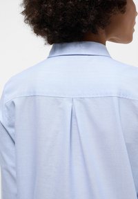 Camicia azzurro chiaro con bottoni, caratterizzata da colletto classico, piega sul retro e una texture liscia e tessuta. Design pulito senza motivi visibili.