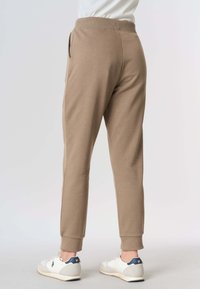 Vista posteriore di una persona che indossa pantaloni jogger color beige a gamba affusolata e sneaker bianche con dettagli blu navy sul tallone, in piedi su pavimento grigio.