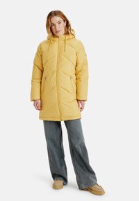 Geel gewatteerde parka met een capuchon, voorzien van chevronstiksels en ritszakken, gecombineerd met een brede spijkerbroek en lichtbruine schoenen.