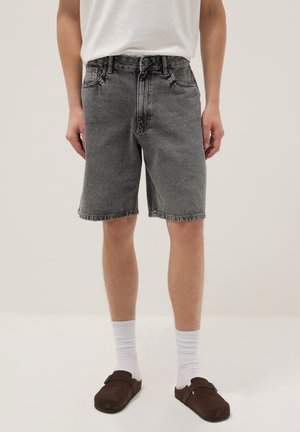 Person trägt graue Jeansshorts, weiße Socken, braune Slipper und ein weißes T-Shirt und steht vor einem schlichten hellen Hintergrund.