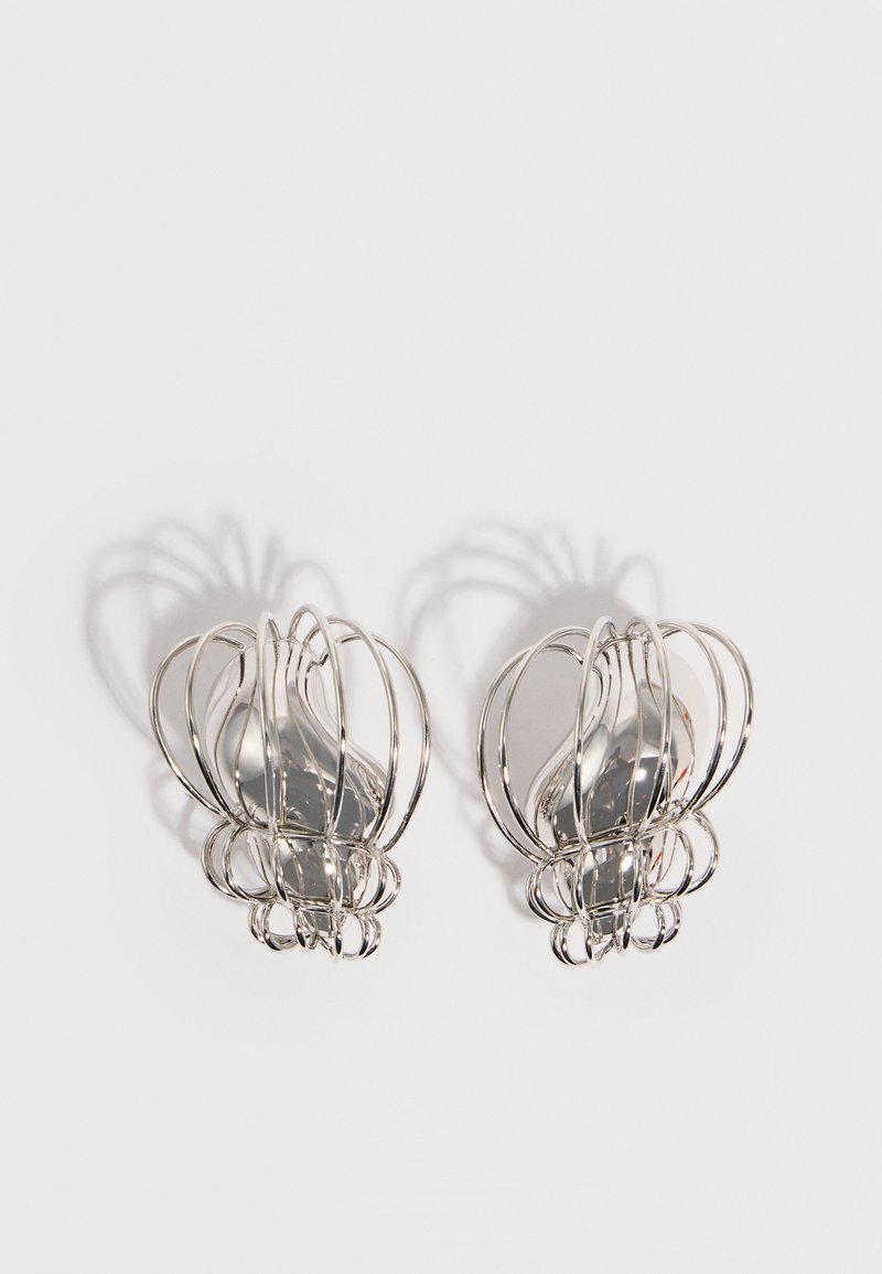 Ζεύγος ασημένιων μεταλλικών σκουλαρικιών με ανοιχτό σχεδιασμό καλυμματός από σύρμα και λείες κεντρικές οβάλ μορφές που ρίχνουν καμπυλωτές σκιές σε λευκό φόντο.