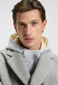 Cappotto grigio in misto lana con design doppiopetto sovrapposto a una felpa leggera grigia. Presenta un rivestimento interno crema e un tessuto testurizzato.