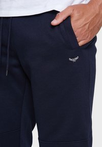 Pantalon de survêtement bleu marine en tissu texturé, doté de poches latérales et d'un petit logo brodé sur la hanche gauche. Taille élastique incluse.