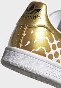 Guld- och vit sneaker med en texturerad läderöverdel, prydd med ett prickmönster, vit gummisula och Adidas-logotyp.