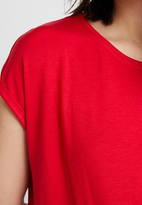 Mujer que lleva una camisa roja de manga corta con puños doblados, mostrando el hombro y parte del cuello y el cabello contra un fondo sencillo.