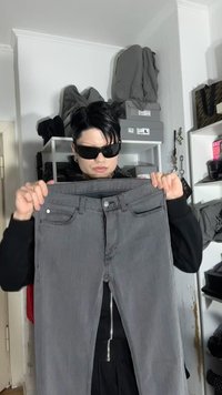 Oseba z očali za sonce drži par sivih kavbojk v prostoru z policami, na katerih so shranjene torbe in škatle.