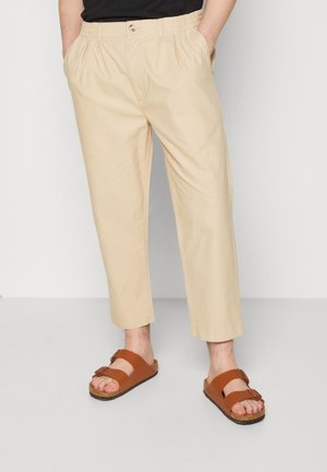 Man die beige korte broek en bruine sandalen met dubbele bandjes draagt, staand met handen in de zakken tegen een effen achtergrond.