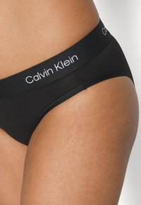 Svart bomulls bikiniundertøy med bred elastisk livlinje som viser "Calvin Klein" i hvit tekst. Glatt tekstur med en tettsittende passform.