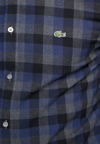 Tela de camisa a cuadros en azul, negro y gris, con costuras visibles, botones y un pequeño logo de cocodrilo verde bordado en el pecho.