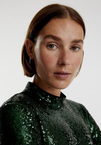 Groene pailletten top met een hoge halslijn en lange mouwen. Heeft dicht op elkaar gepakte ronde pailletten die een textuur en reflecterend oppervlak creëren.