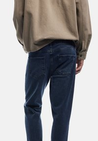 Donkerblauwe jeans van denim, met twee achterzakken en een licht taps toelopende pijp. Een losse beige top is gedeeltelijk zichtbaar erboven.