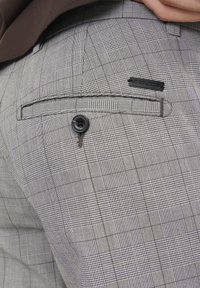 Detailní záběr na šedé kostkované kalhoty s knoflíkem na zadní kapse a černou visačkou Jack & Jones, ruka spočívající na pase.