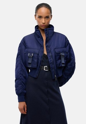 Mujer que lleva una chaqueta acolchada corta azul marino con grandes bolsillos sobre un vestido oscuro abotonado con un cinturón negro, de pie frente a un fondo liso.