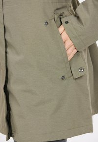 Olijfgroene jas van textuurstof, voorzien van een zijzak met knoopsluiting en een gladde zoom. Het ontwerp bevat verstevigde naden.