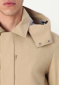 Beige vandtæt jakke med høj krave, knapdetaljer og frontlynlås. Har en snorejustering og en glat tekstur.