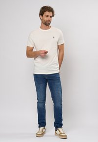 Beige kortermet t-skjorte med en liten logo på brystet, sammen med blå denimjeans og tan- og hvite sneakers.