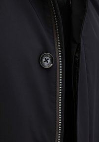 Sort jakke med en glat overflade, der har en sølv lynlås og en rund sort knap med blomsterdesign.