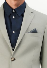 Ανοιχτού γκρι σακάκι με ένα κουμπί, συνδυασμένο με σκούρο ναυτικό πουκάμισο και σκούρο μαντήλι στην τσέπη του στήθους. Λείο υλικό.