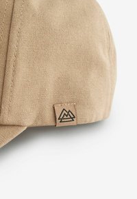 Beige keps i texturerad tyg, med en liten märkesetikett med en berglogga sydd på sidan.