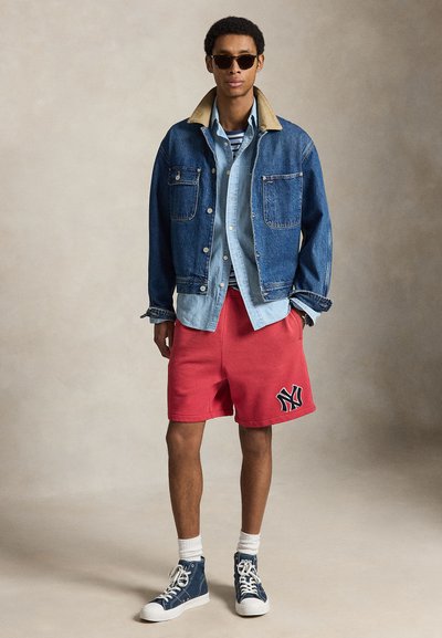 Homme portant des lunettes de soleil, plusieurs vestes en jean superposées, un short rouge avec un logo, des chaussettes blanches et des baskets montantes bleues, debout devant un fond uni.