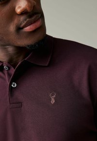 Burgundy polo tričko vyrobené z textúrovanej látky, s golierom, dvoma gombíkmi a vyšitým logom jelena na ľavej hrudi.