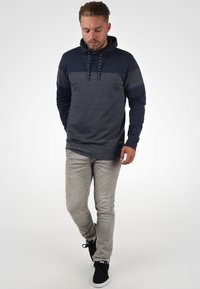 Felpa con cappuccio navy con pannello grigio, coulisse e maniche lunghe. Abbinata a jeans grigi chiari e sneaker nere.