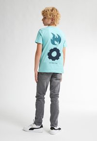 Camiseta de algodón turquesa con gráficos de llamas y engranajes en navy en la parte posterior, combinada con jeans de mezclilla grises y zapatillas blancas con acentos negros.
