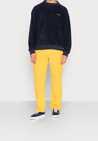 Felpa in pile blu navy con polsini a coste e scollo rotondo, abbinata a pantaloni in corduroy gialli. Il modello indossa sneakers blu navy con suole bianche.