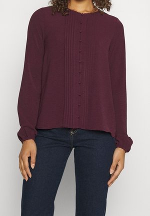 Kvinde iført en blommefarvet bluse med lange ærmer med stofovertrukne knapper og plisseringer foran, kombineret med mørkeblå jeans.