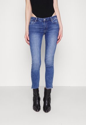 Slim-fit blå jeans båret med sorte ankelstøvler og en sort top, vist fra talje til fødder mod en ensfarvet hvid baggrund.